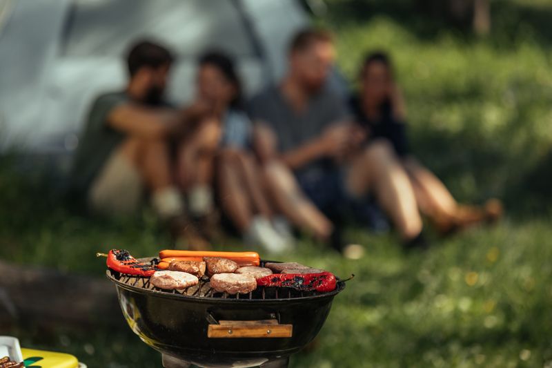 A BBQ cooking food outdoors with a person and a tent.